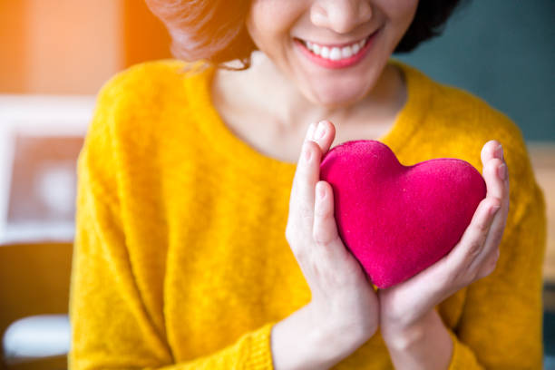 close up of woman hands in yellow sweater holding pink heart. people, age, family, love, valentine and health care concept